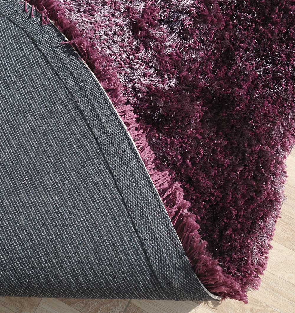 A close-up of the RAMSHA HOME Shaggy Rug for Living Room - SG 019 in deep plum, showing its plush pile and one corner lifted to reveal the black, textured underside on a wooden floor.