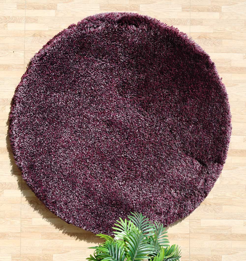 The RAMSHA HOME Shaggy Rug for Living Room - SG 019, a round dark purple plush rug, lies on a light wood floor with a green potted plant partly seen at the bottom edge of the image.