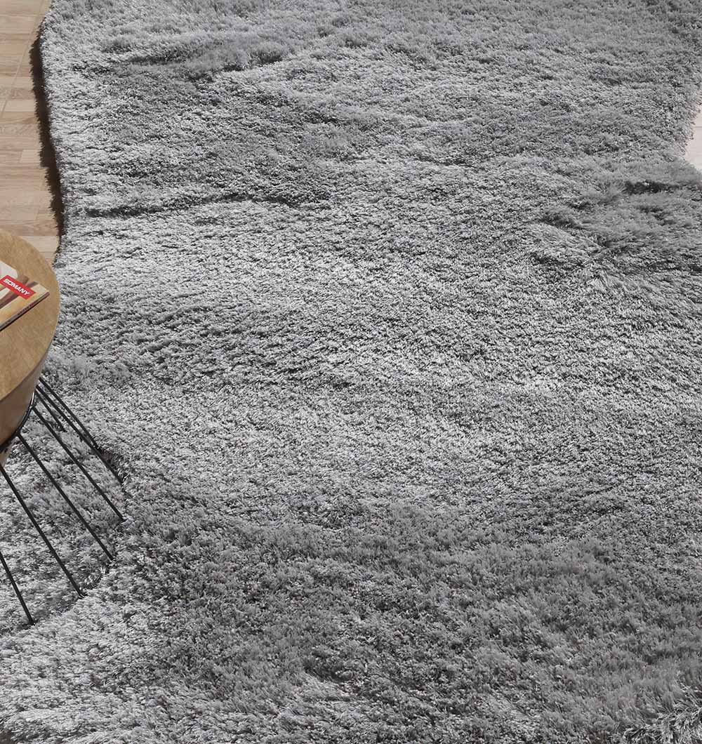 A close-up of the RAMSHA HOME Shaggy Shape Rug for Living Room - SG 020 (175x300 cm), plush and grey with an irregular shape, lies on a wooden floor; part of a round tan chair with metal legs appears in the upper left corner.