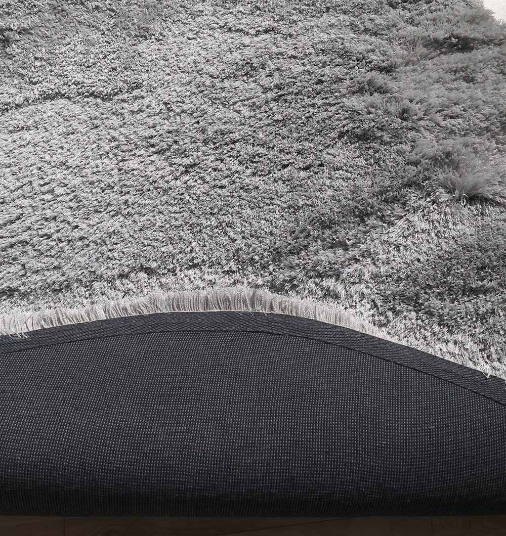 Close-up of the RAMSHA HOME Shaggy Shape Rug for Living Room - SG 020 (175x300 cm), showing its soft grey plush surface, one corner flipped to reveal a dark non-slip backing, and fringe detail along the edge.