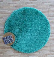 The RAMSHA HOME Turquoise Fluffy Shaggy Round Shape Area Rug SG 021 lies on a light wood floor, with a small round wooden table and a patterned book or magazine placed partly on the rug’s edge.