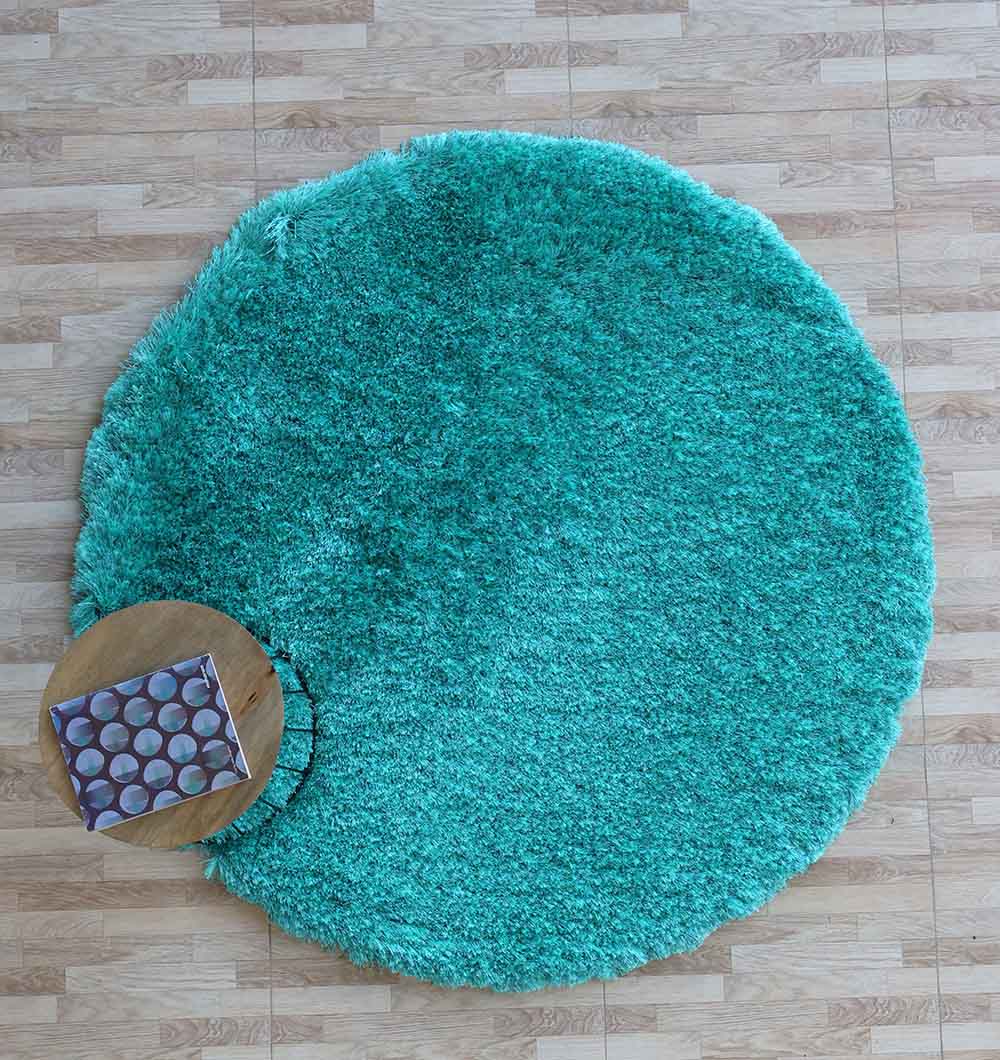 The RAMSHA HOME Turquoise Fluffy Shaggy Round Shape Area Rug SG 021 lies on a light wood floor, with a small round wooden table and a patterned book or magazine placed partly on the rug’s edge.