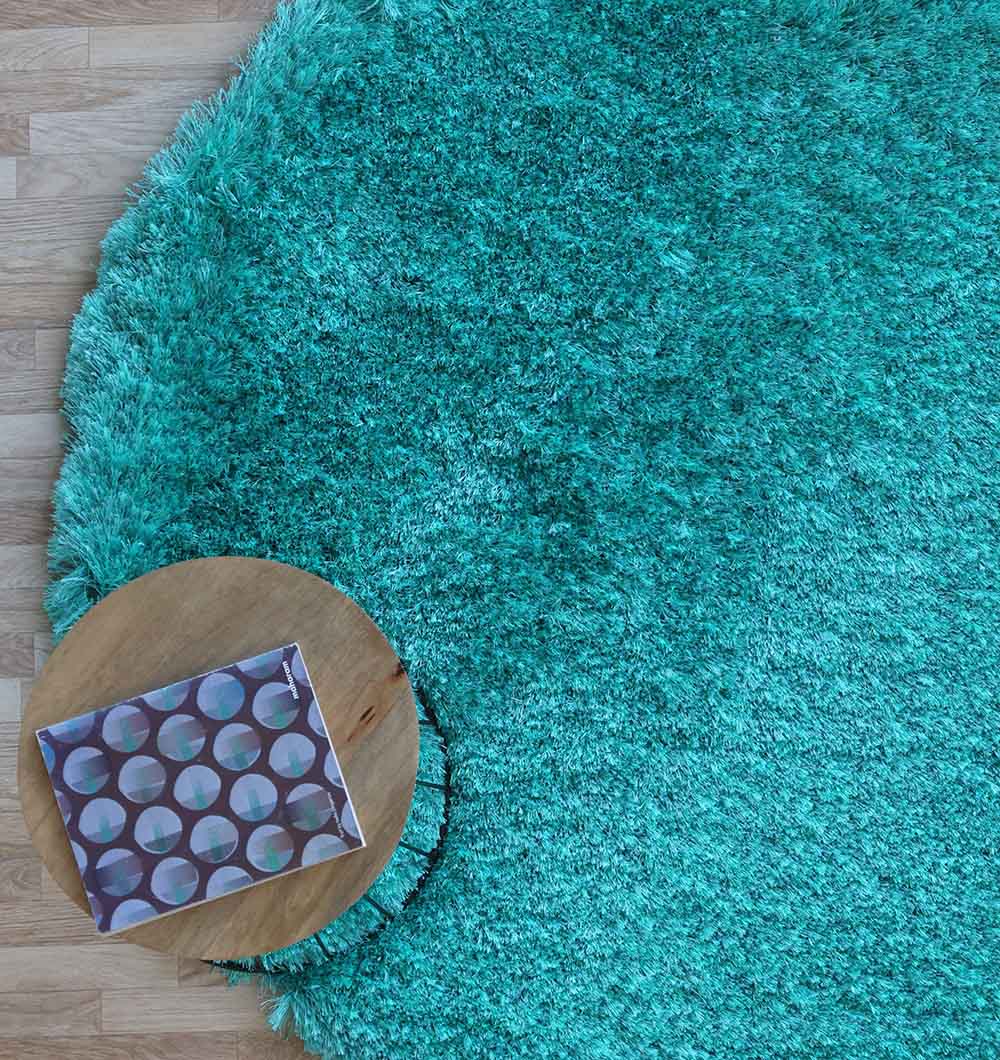 A RAMSHA HOME Turquoise Fluffy Shaggy Round Shape Area Rug SG 021 lies on a wooden floor with a small round wooden table on top. A closed notebook with a geometric circle pattern rests on the table.