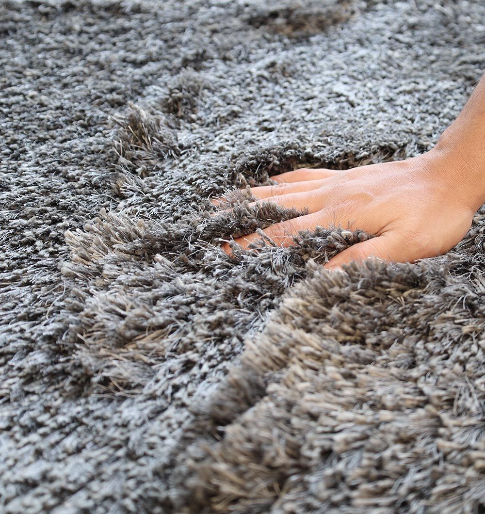 A hand presses into the plush fibers of the RAMSHA HOME Shaggy Shape Rug for Living Room - SG 022 (200x300 cm), making the soft, gray shaggy texture stand out and emphasizing its unique irregular shape.