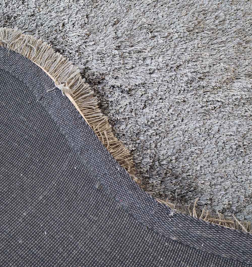 A close-up of the edge of the RAMSHA HOME Shaggy Shape Rug for Living Room - SG 022 (200x300 cm), showing its plush gray texture, beige fringe, and dark, textured underside partially flipped over.