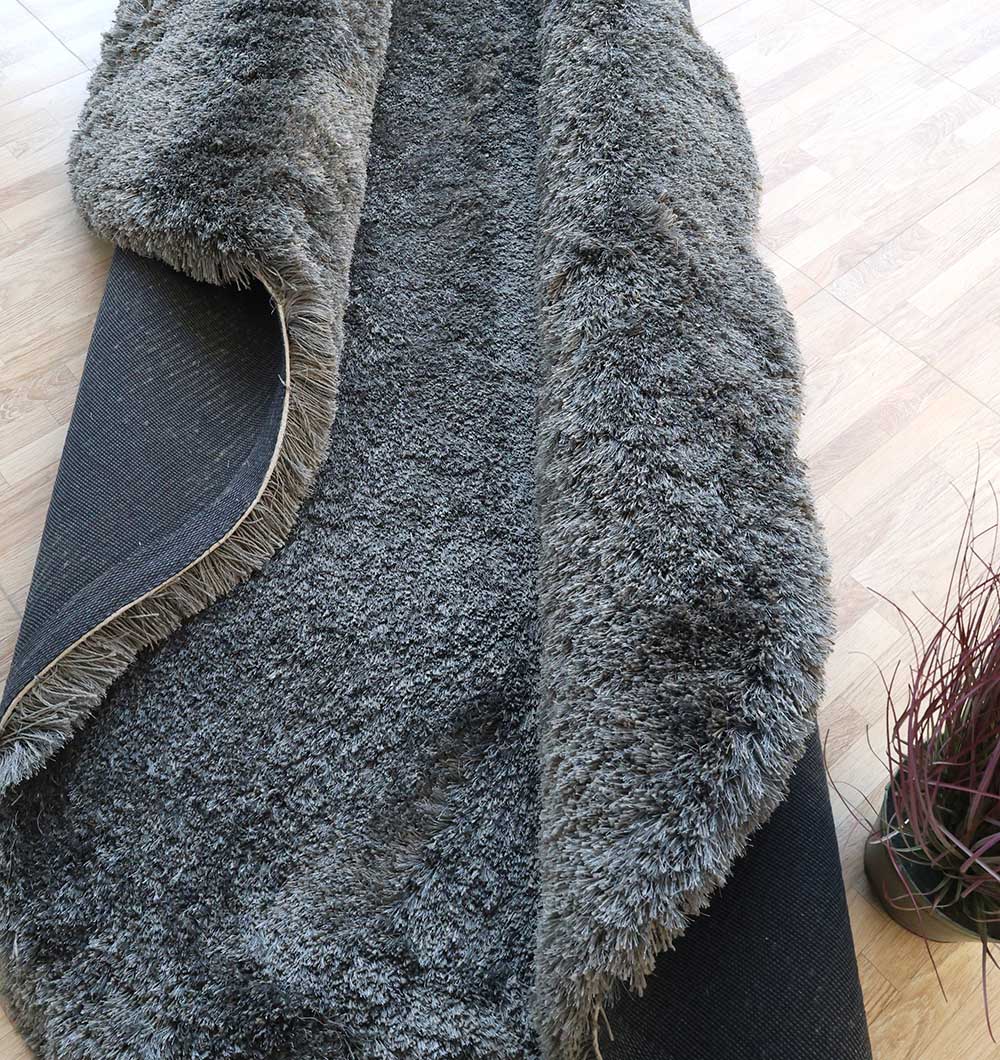 The RAMSHA HOME Shaggy Shape Rug for Living Room - SG 022 (200x300 cm), in gray with an irregular form and plush texture, is partly rolled up on a light wooden floor beside a small plant with thin, dark red leaves.