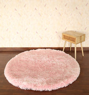 The Light Pink Color Home Decor Round Shaggy Rug - SG 02 sits on a dark wooden floor beside a small light wood side table, set against a beige patterned wallpapered wall.