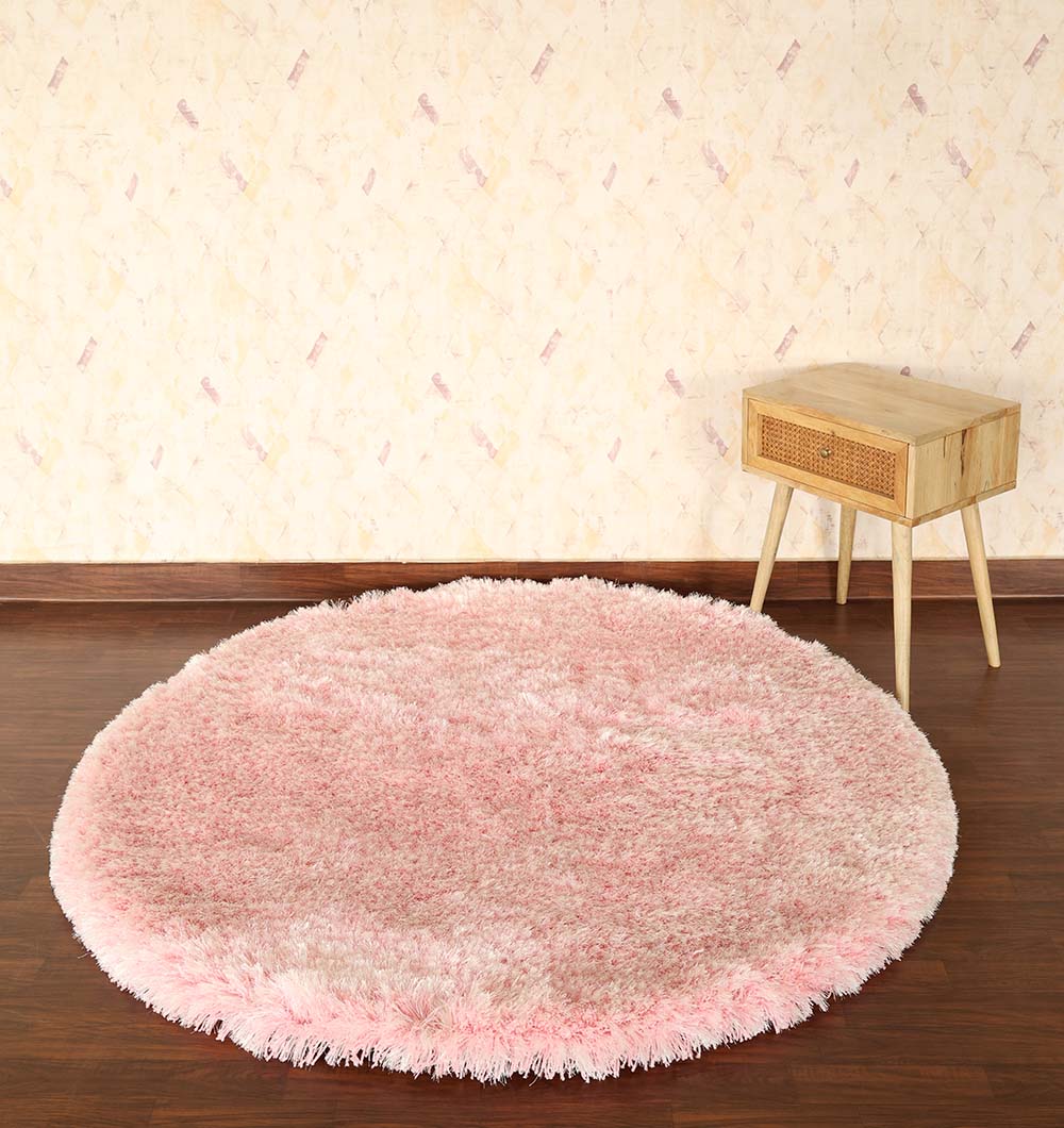 The Light Pink Color Home Decor Round Shaggy Rug - SG 02 sits on a dark wooden floor beside a small light wood side table, set against a beige patterned wallpapered wall.