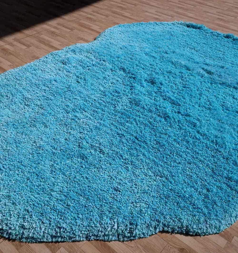 The RAMSHA HOME Shaggy Rug for Living Room - SG 023 (200x300 cm), in turquoise with an irregular shape, lies on a wooden floor. Its soft, plush texture resembles a pond’s outline, and sunlight casts gentle shadows across the surface.