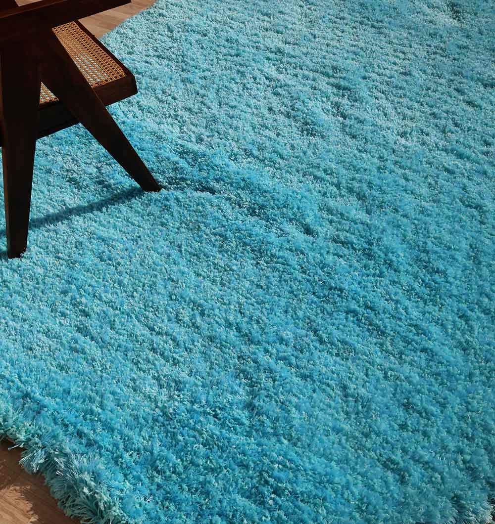 A close-up of the RAMSHA HOME Shaggy Rug for Living Room - SG 023 (200x300 cm), featuring a plush blue shag texture and irregular shape on a wooden floor, with part of a dark wooden chair in the upper left corner.