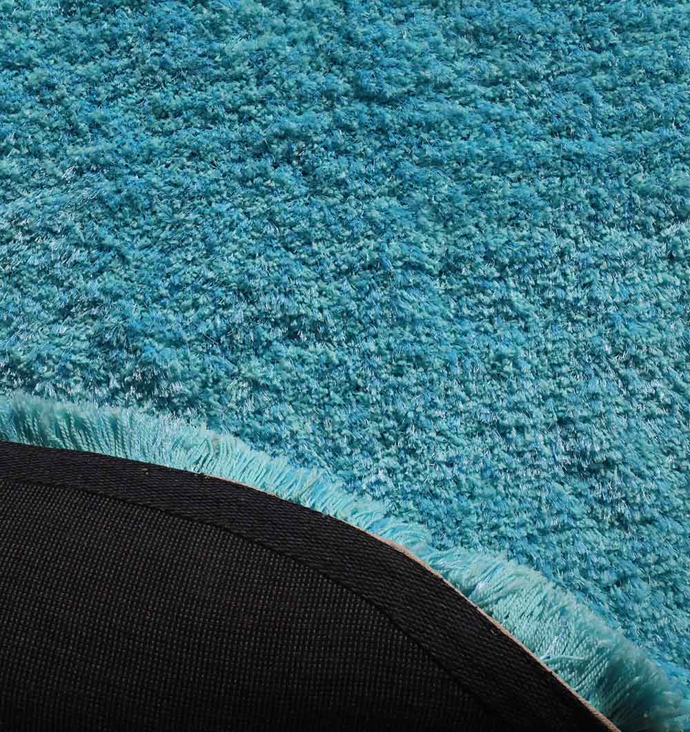 Close-up of the RAMSHA HOME Shaggy Rug for Living Room - SG 023 (200x300 cm) in bright teal blue, showing its soft textured surface, lifted corner revealing black backing, and fringe along the edge.