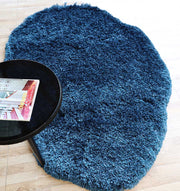 The RAMSHA HOME Blue Color Home Decor Shaggy Rug - SG 024, with its soft, irregular shape, rests on a wooden floor beside a round black table holding a magazine.