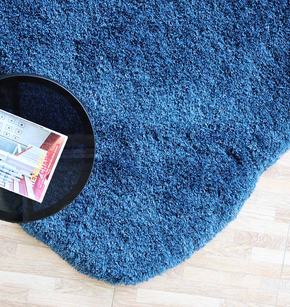 The RAMSHA HOME Blue Color Home Decor Shaggy Rug - SG 024, featuring a soft navy blue shag and irregular round shape, rests on a light wooden floor beside a small black table topped with a colorful magazine.