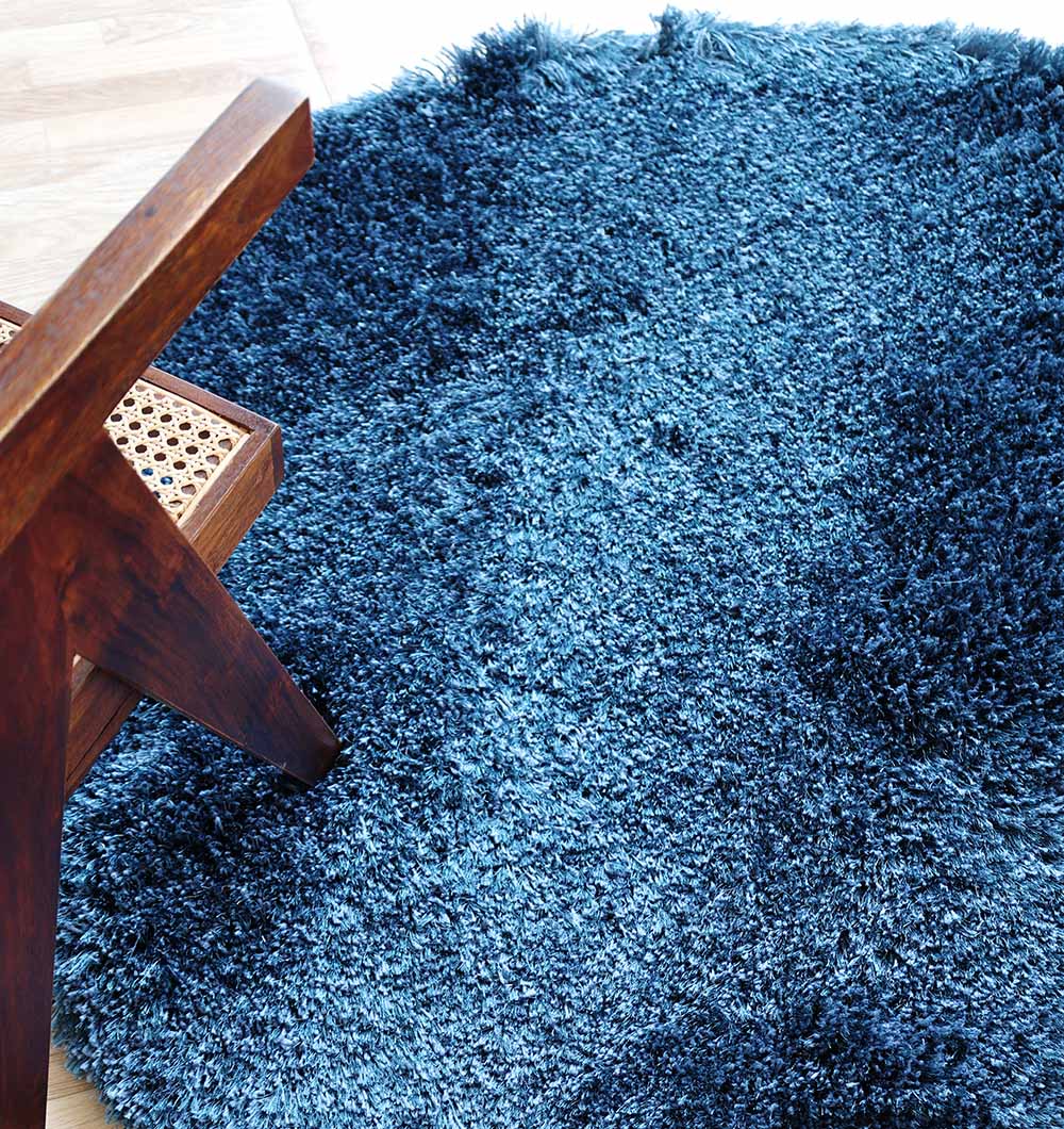 The RAMSHA HOME Blue Color Home Decor Shaggy Rug - SG 024, featuring a soft shaggy texture and irregular round shape, is shown next to a wooden chair with a woven seat on a light-colored floor.