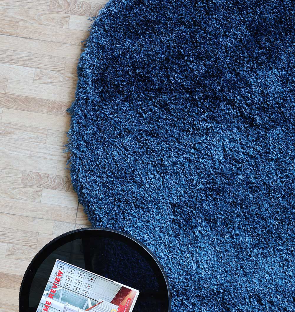 The RAMSHA HOME Blue Color Home Decor Shaggy Rug - SG 024, round and textured in navy blue, lies on a light wood floor with a small black table holding magazines at its edge.