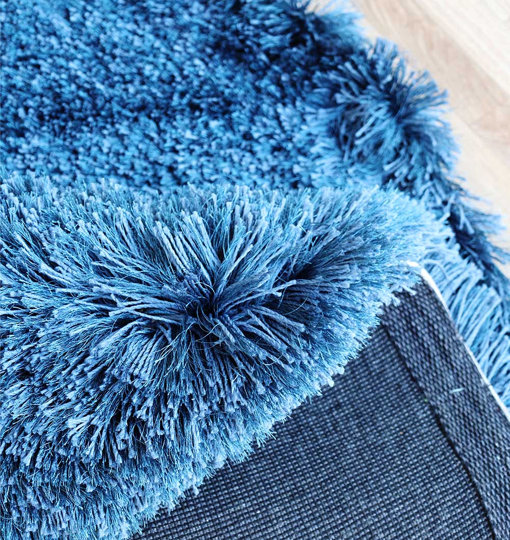 Close-up of RAMSHA HOME's Blue Color Home Decor Shaggy Rug - SG 024 with long, fluffy navy fibers, corner lifted to reveal the textured underside. This cozy rug adds warmth to a light wooden floor.