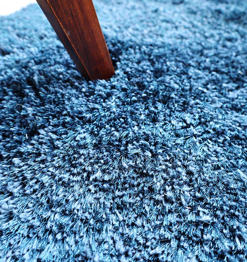 Close-up of RAMSHA HOME Blue Color Home Decor Shaggy Rug - SG 024, featuring thick, soft navy blue fibers. A wooden furniture leg rests gently on the plush surface in the upper left corner.