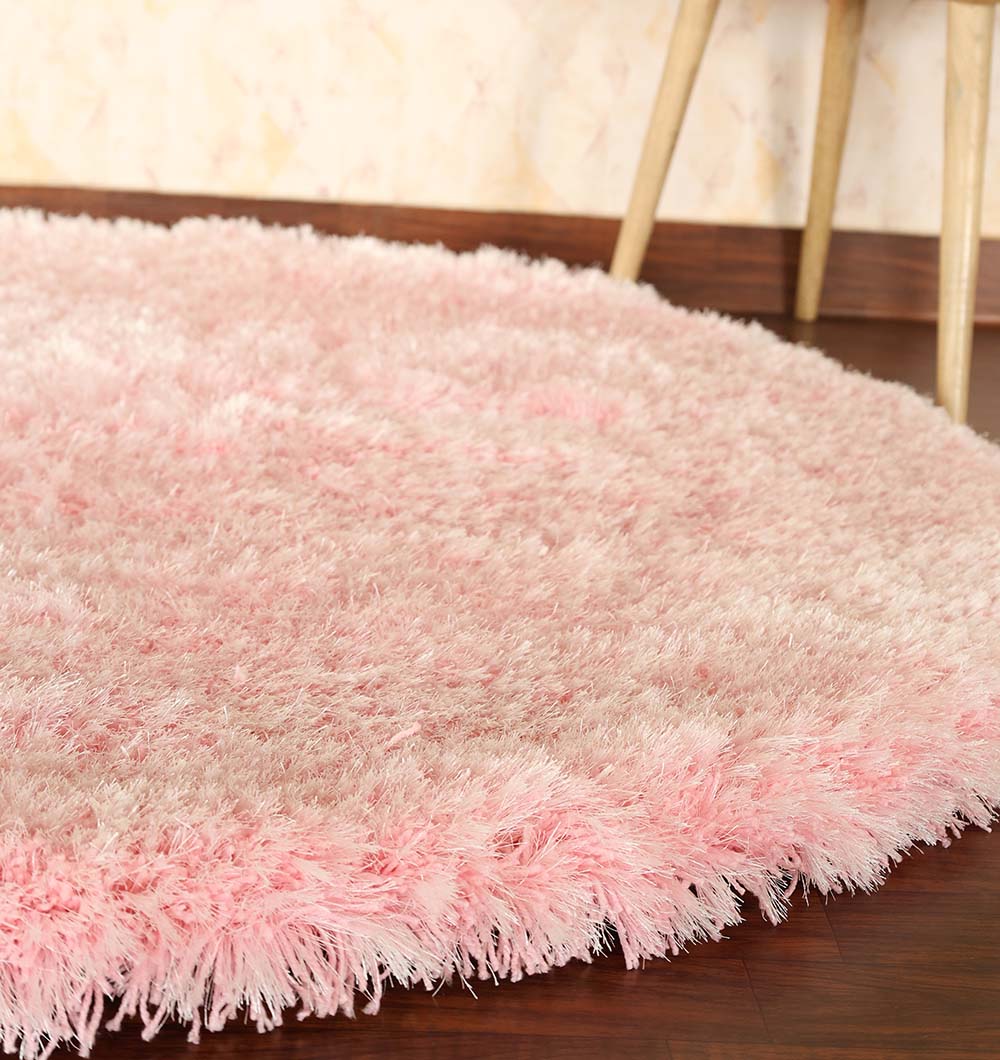 The Light Pink Color Home Decor Round Shaggy Rug - SG 02 adds a soft, fluffy touch to a dark wooden floor beside a light wall and the legs of a wooden table or stool.