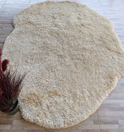 The RAMSHA HOME Shaggy Rug for Living Room (SG 026), a large plush ivory rug with an irregular shape, sits on a light wood floor. A plant with burgundy leaves in a black pot is partially visible in the lower left corner.
