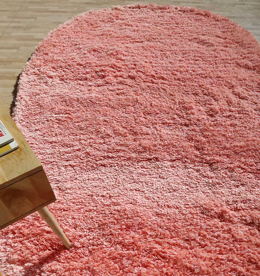 The RAMSHA HOME Shaggy Shape Rug for Living Room - SG 027-A (200x300 cm), a large oval pink shag rug, lies on a light wooden floor with the corner of a wooden table and book visible at the bottom left.