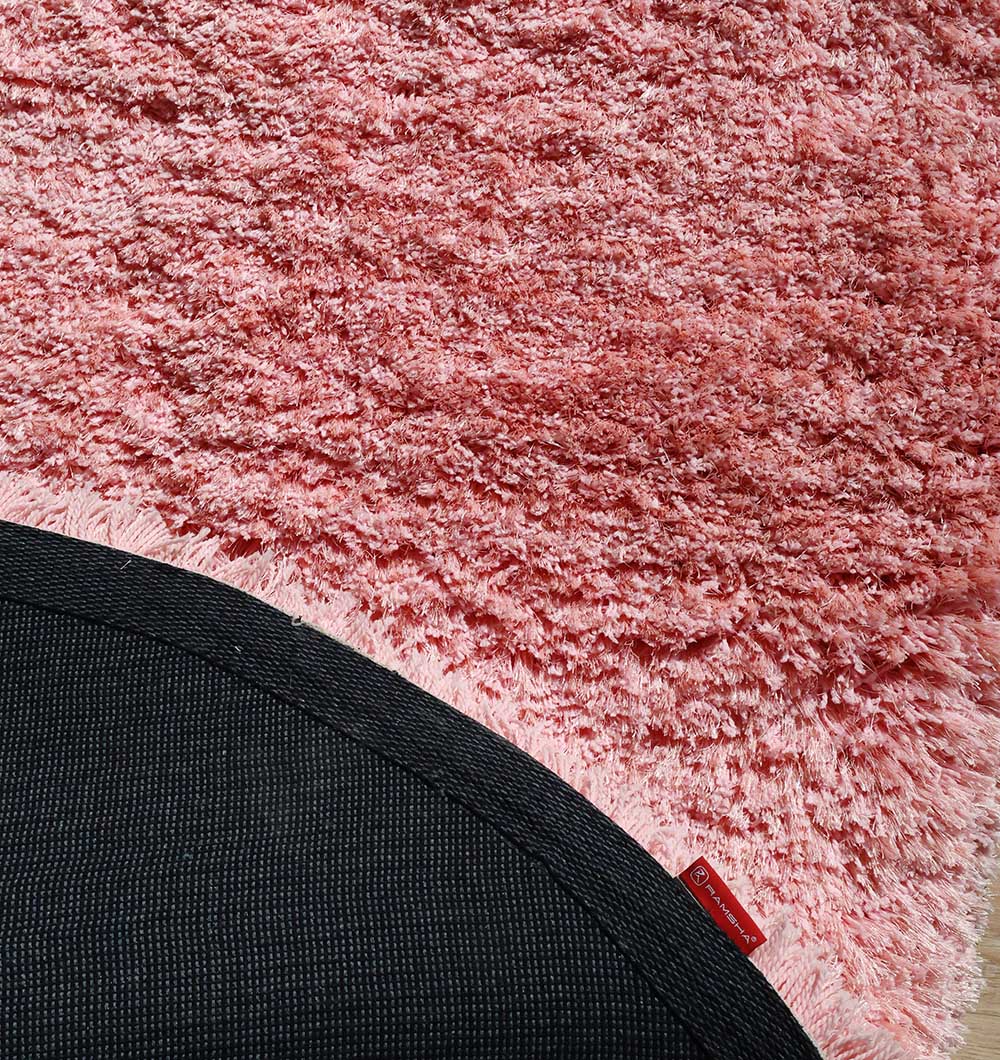 A close-up of the RAMSHA HOME Shaggy Shape Rug for Living Room - SG 027-A (200x300 cm), pink with an irregular shape, partly covered by a rounded black mat. Sunlight highlights its plush texture and a small red fabric tag near the edge.