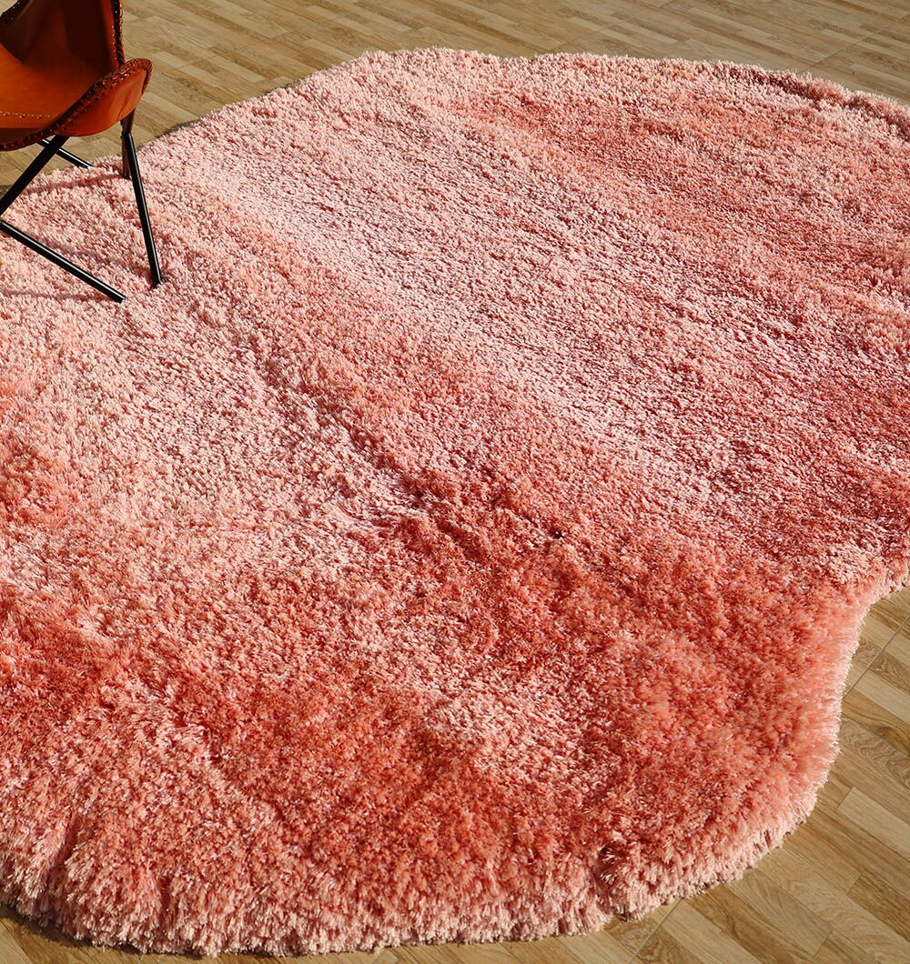 The RAMSHA HOME Shaggy Shape Rug for Living Room - SG 027-B (200x300 cm) features a plush pink shaggy, abstract-shaped design with a textured surface on a light wooden floor; an orange chair is visible at one corner.