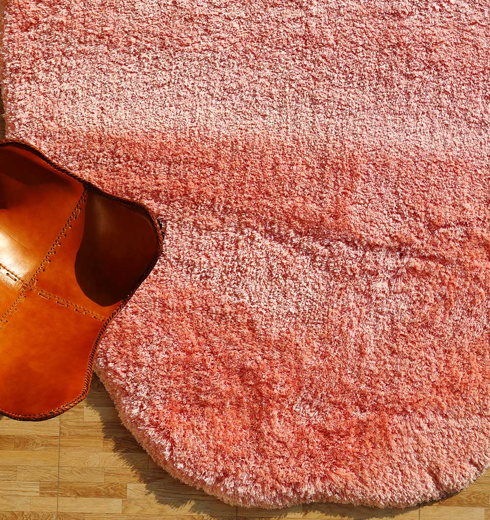 The RAMSHA HOME Shaggy Shape Rug for Living Room - SG 027-B (200x300 cm), in pink with an irregular edge, is partially covered by a tan leather chair on a light wooden floor.