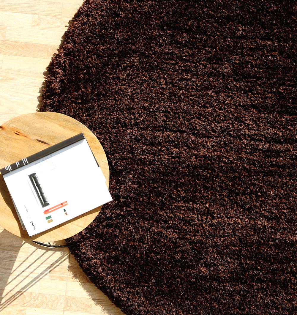 A Dark Brown High-Density Shaggy Rug - SG 028 by RAMSHA HOME sits on a light wooden floor, with a small wooden side table holding an open book or magazine at the rug’s edge.