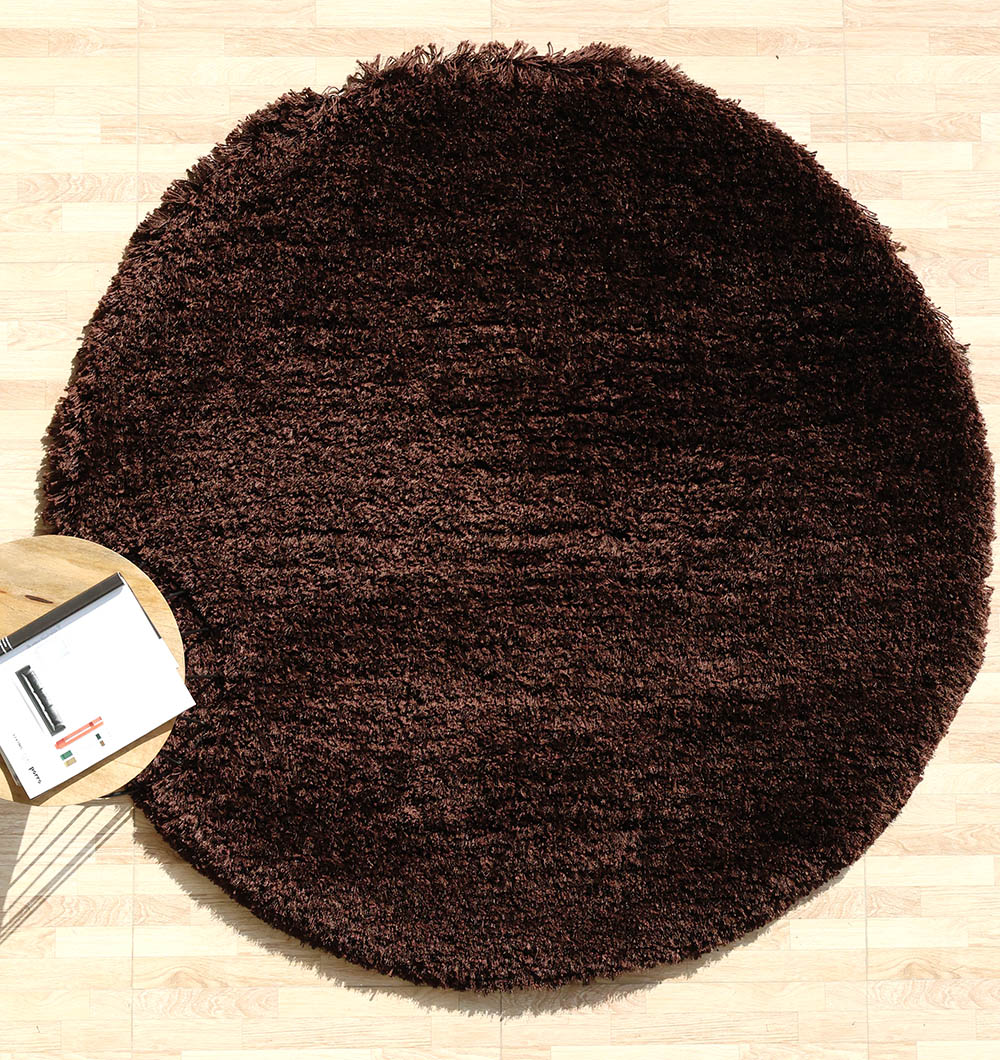 The RAMSHA HOME Dark Brown High-Density Shaggy Rug - SG 028 adds coziness atop a light wood floor, paired with a small round wooden table holding an open magazine and pen in the lower left corner.