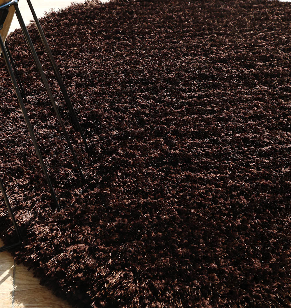 A close-up of the RAMSHA HOME Dark Brown High-Density Shaggy Rug - SG 028, featuring a plush texture and unique shape, is shown on a light wood floor with black metal furniture legs visible on the left.