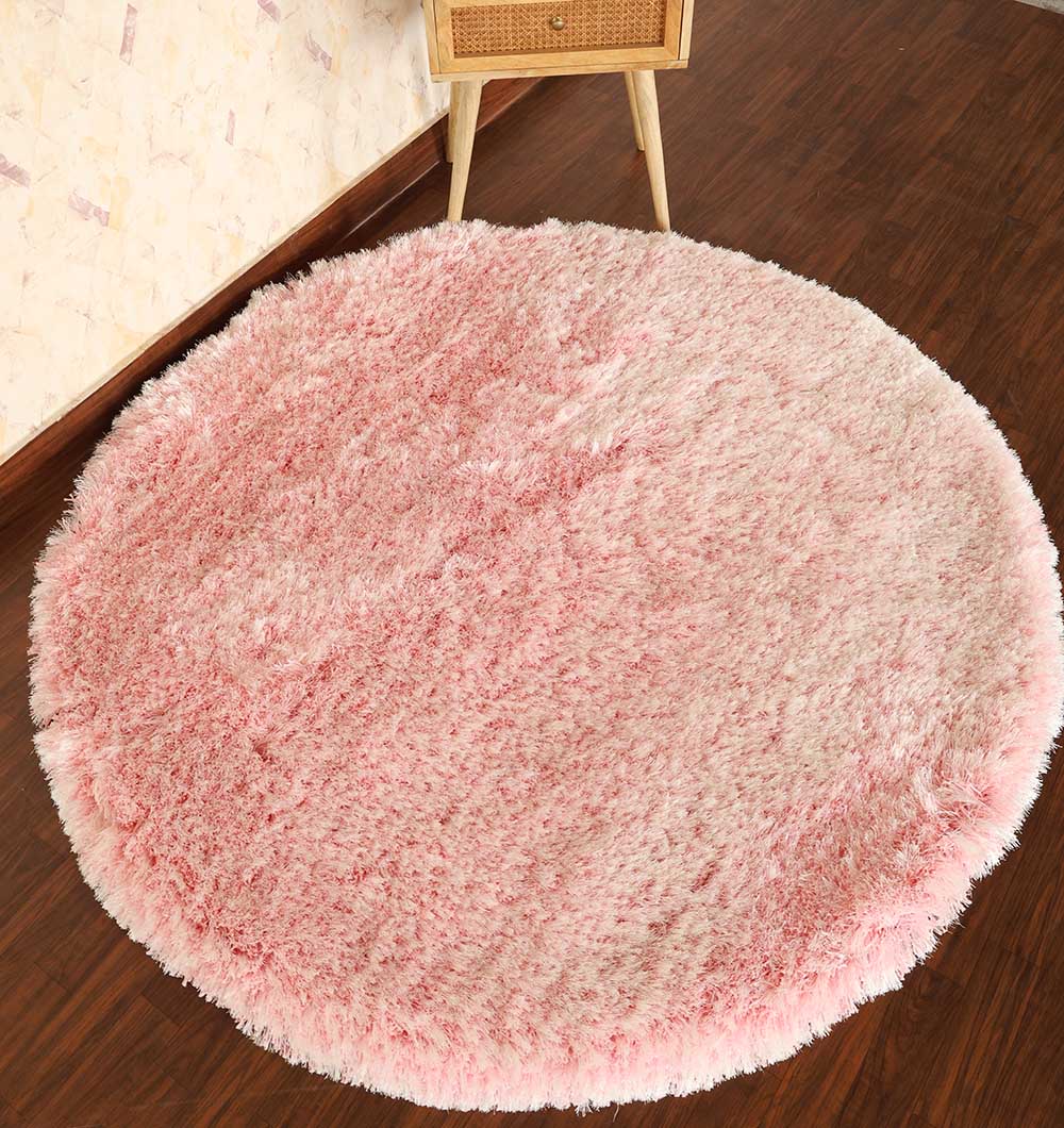 The Light Pink Color Home Decor Round Shaggy Rug - SG 02 adds a cozy touch on a dark wooden floor beside a corner with light-patterned wallpaper and a small wooden side table.