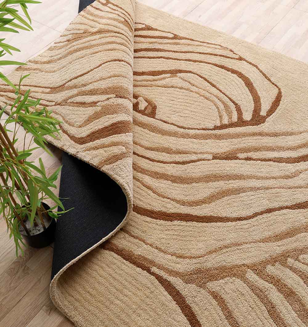 The RAMSHA HOME Rectangle Multi Color Shaggy Rug for Living Room (SG 031, 200x300 cm) features a beige woodgrain pattern, black non-slip backing, and is shown partially rolled on a light wood floor beside a green potted plant.