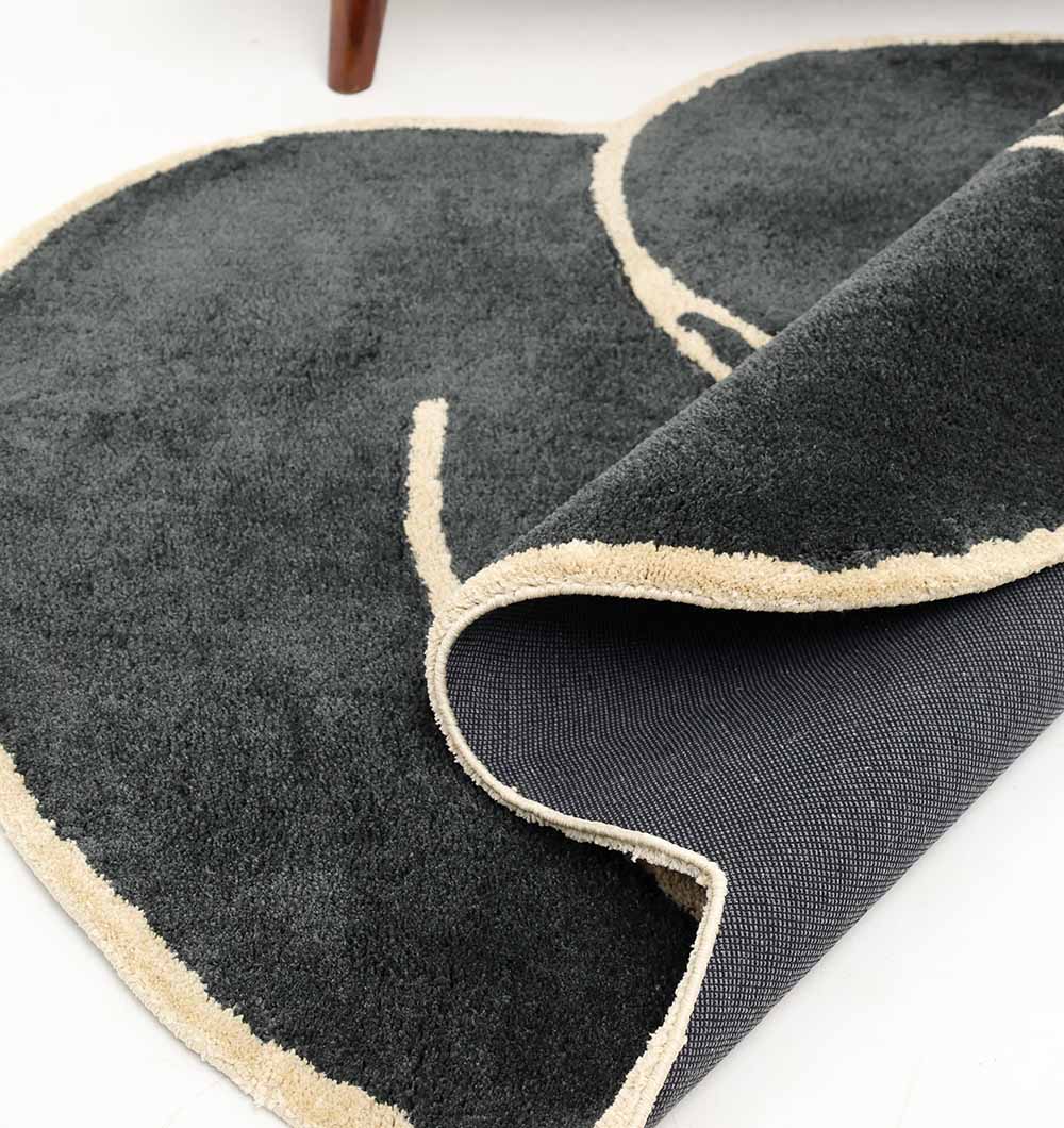 A close-up of the RAMSHA HOME Black & Gold Color Shaped Shaggy Rug for Living Room (SG 034, 160x230 cm), partially folded to reveal its textured backing, is shown on a white floor.