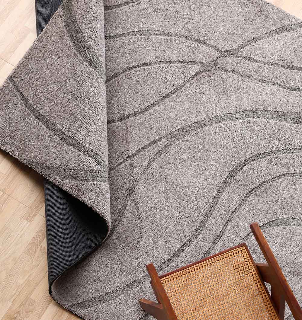 The Shaggy Rug for Living Room - SG 039 (200x300 cm) by RAMSHA HOME features a light gray, wavy abstract pattern with a plush pile shown partially flipped to reveal its dark underside, beside a wooden chair on a light wood floor.