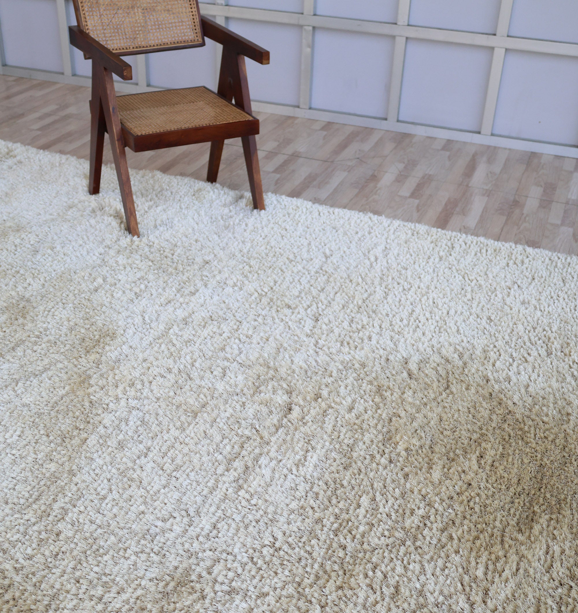 A wooden chair with a woven seat and backrest sits on the RAMSHA HOME Modern Ultra Soft Shaggy Carpet for Bedroom - SG 041 in cozy beige, atop light wood floors, with a grid-patterned wall completing this stylish modern interior.