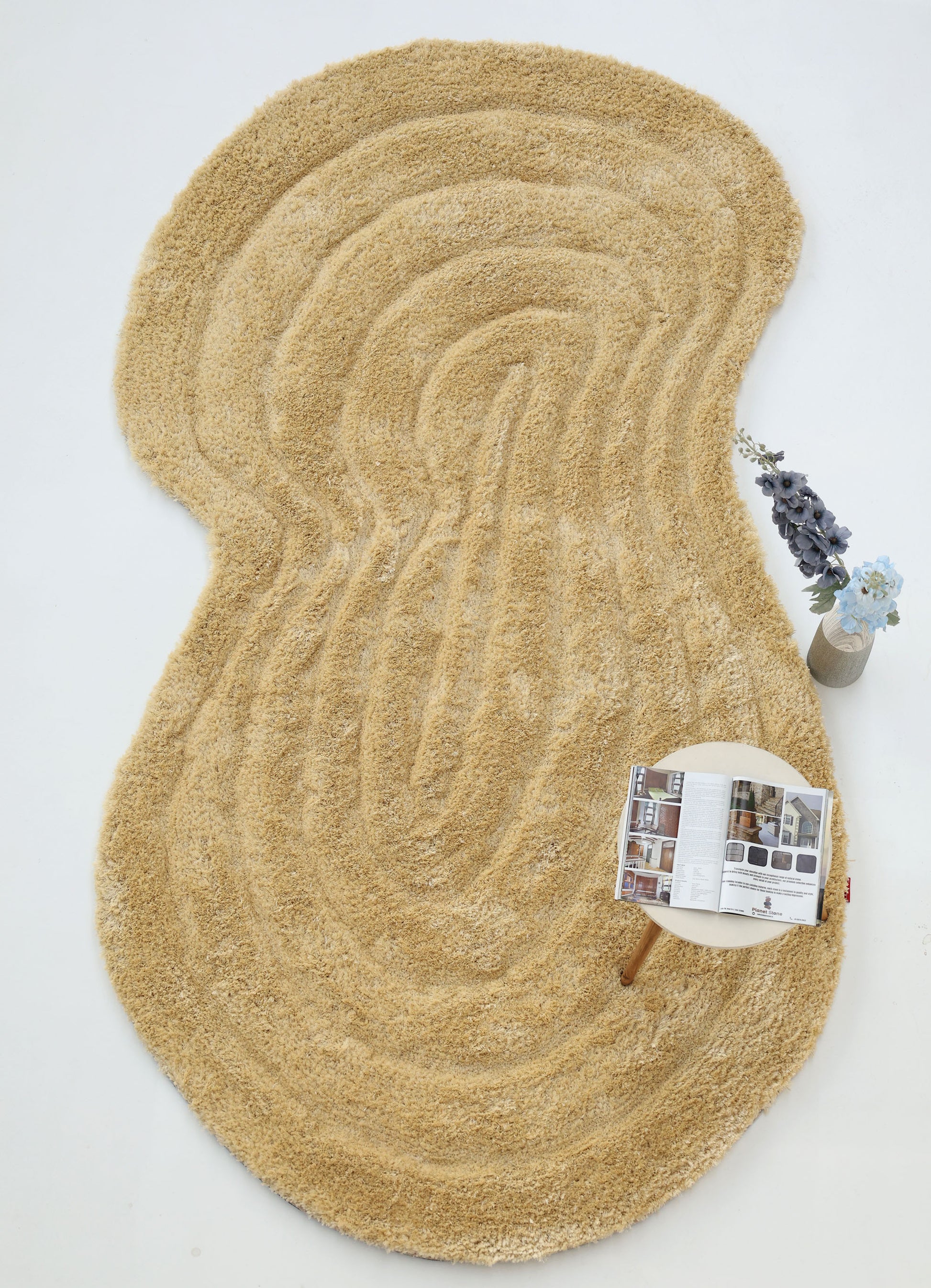 The RAMSHA HOME Gold Irregular Shaped Shaggy Rug (SG 041 3D) sits on a white floor with an open magazine on a small round table and a vase of blue and white flowers nearby.