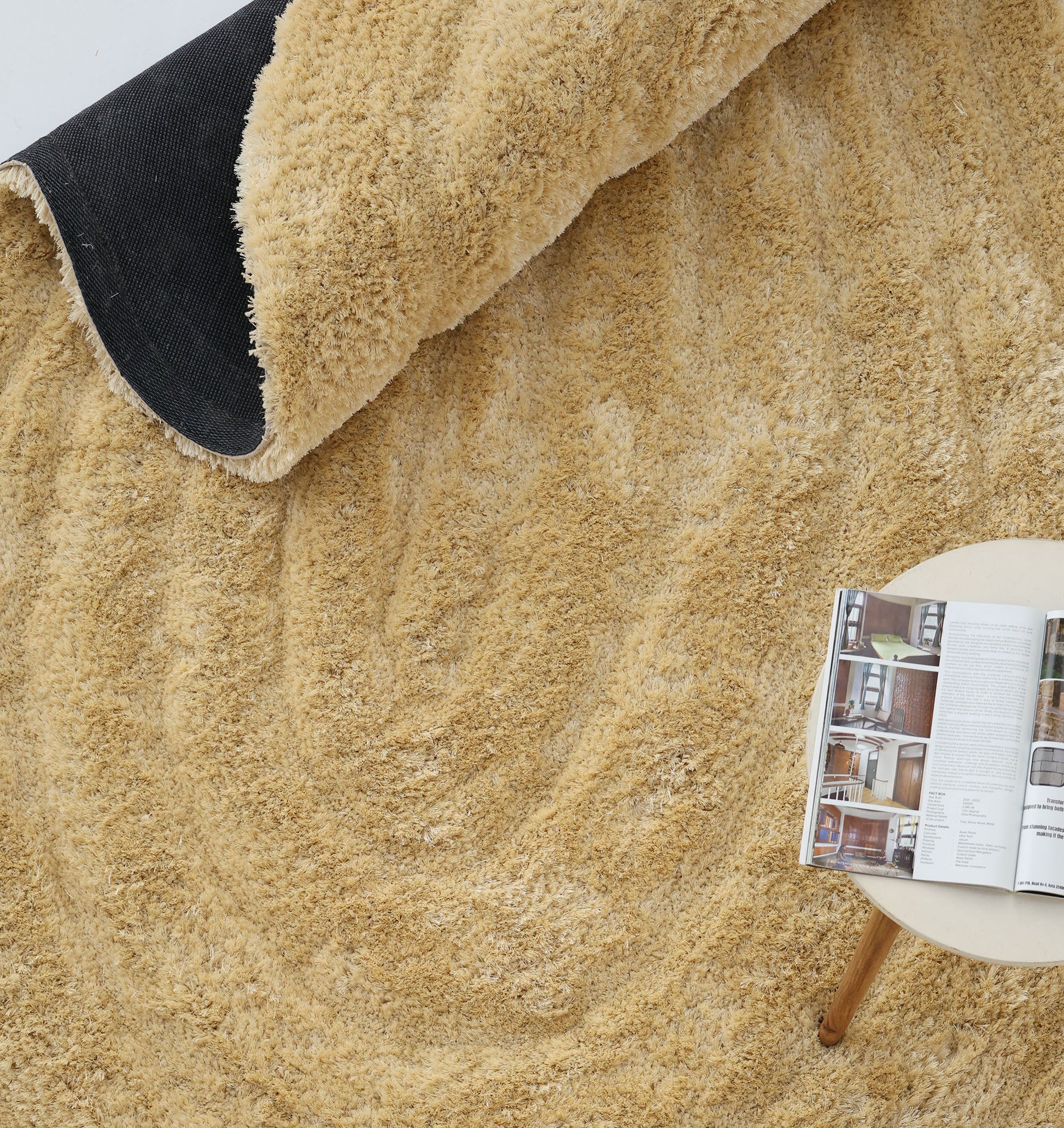 The RAMSHA HOME Gold irregular Shaped Home Decor Shaggy Rug - SG 041 3D is partially rolled up to reveal its black underside, beside a round light table displaying an open magazine with home interior photos.
