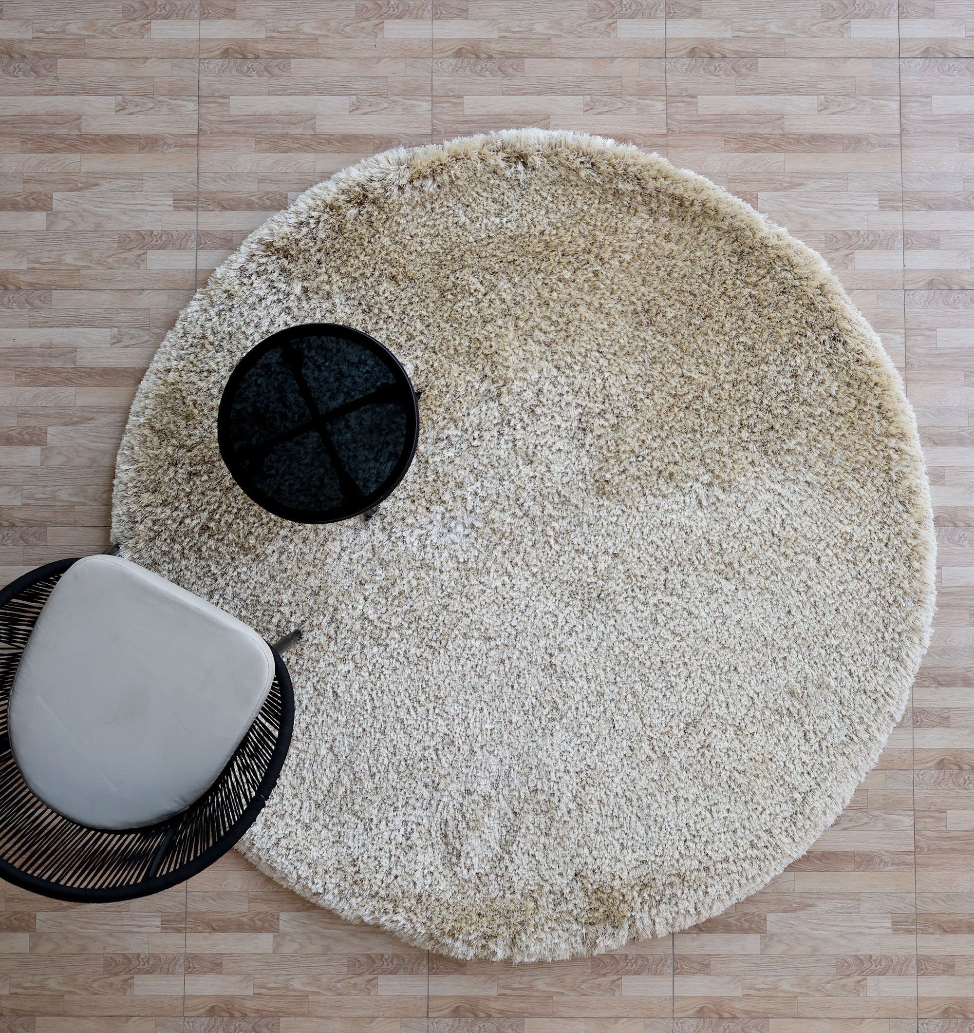 Top view of the RAMSHA HOME Modern Ultra Soft Shaggy Carpet for Bedroom - SG 041 in beige, placed on tiled flooring with a small black round table and part of a white curved chair—ideal for modern interiors.