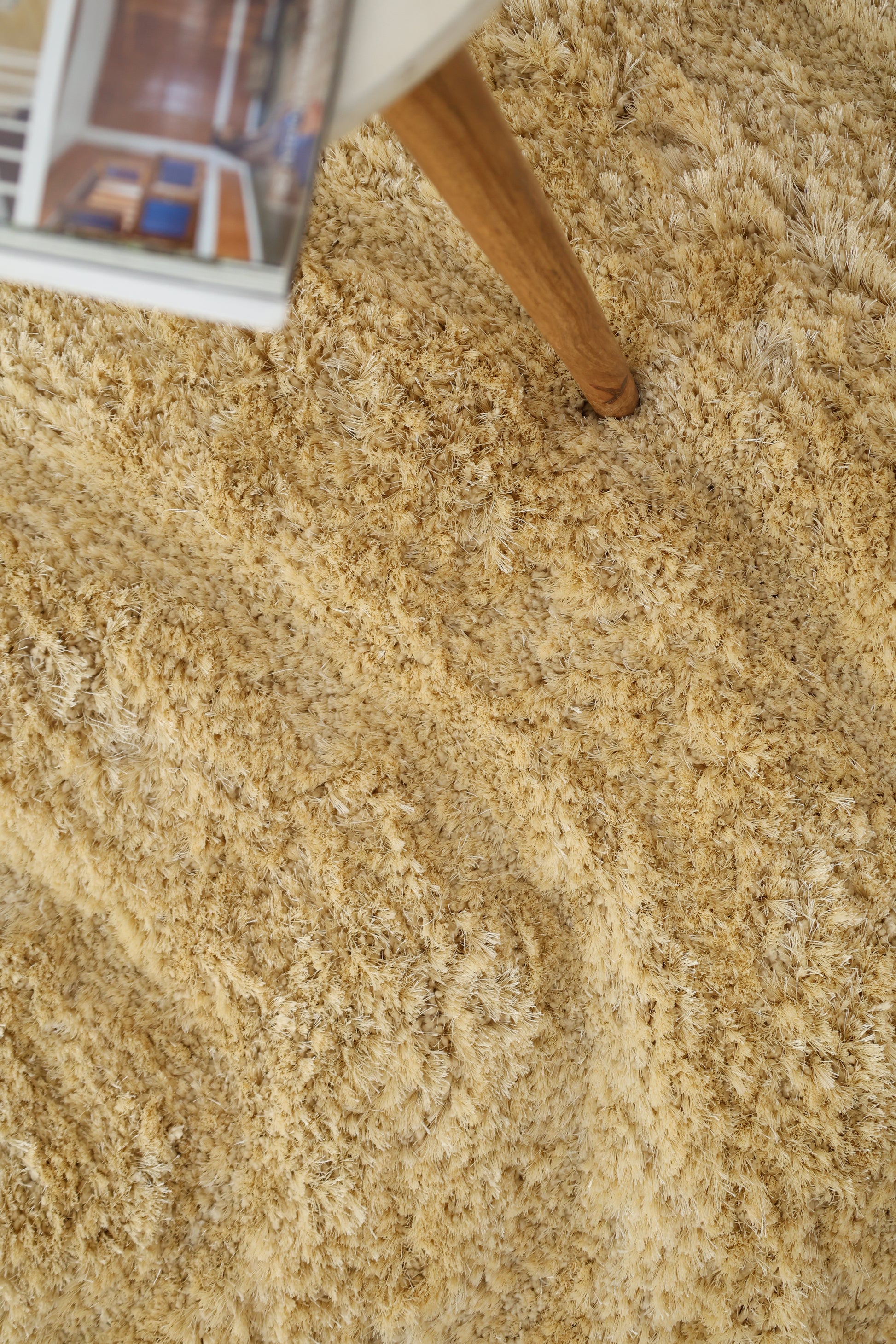 A close-up of the RAMSHA HOME Gold Irregular Shaped Home Decor Shaggy Rug - SG 041 3D in beige, featuring a wooden table leg and the edge of an open magazine in the top left corner.