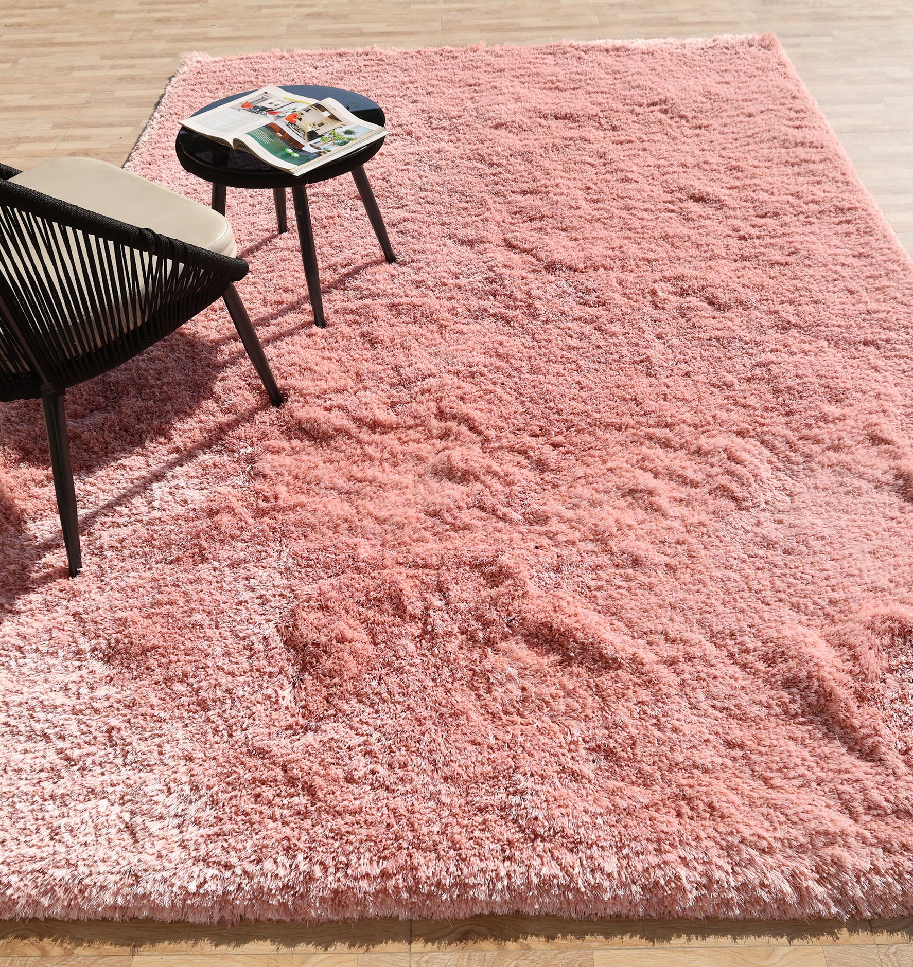 The RAMSHA HOME Modern Ultra Soft Pink Shaggy Carpet for Bedroom (SG 042) highlights a cozy corner with a sleek black chair and round table on a wooden floor—perfect for stylish, modern interiors.