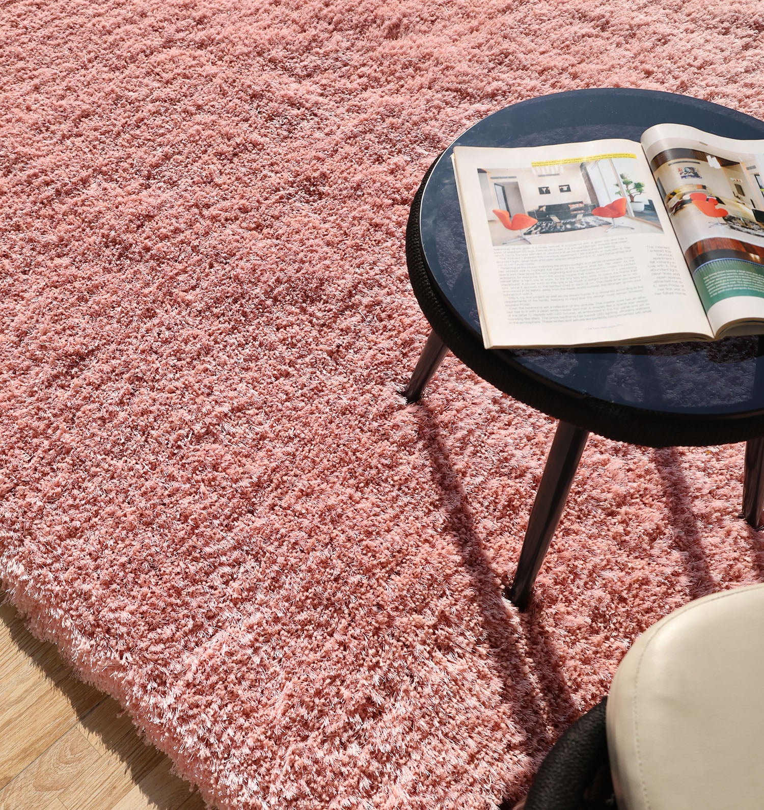 A round black table with an open magazine sits on the RAMSHA HOME Modern Ultra Soft Pink Color Shaggy Carpet for Bedroom (SG 042). Sunlight highlights the plush rug, with part of a beige chair visible in the foreground.
