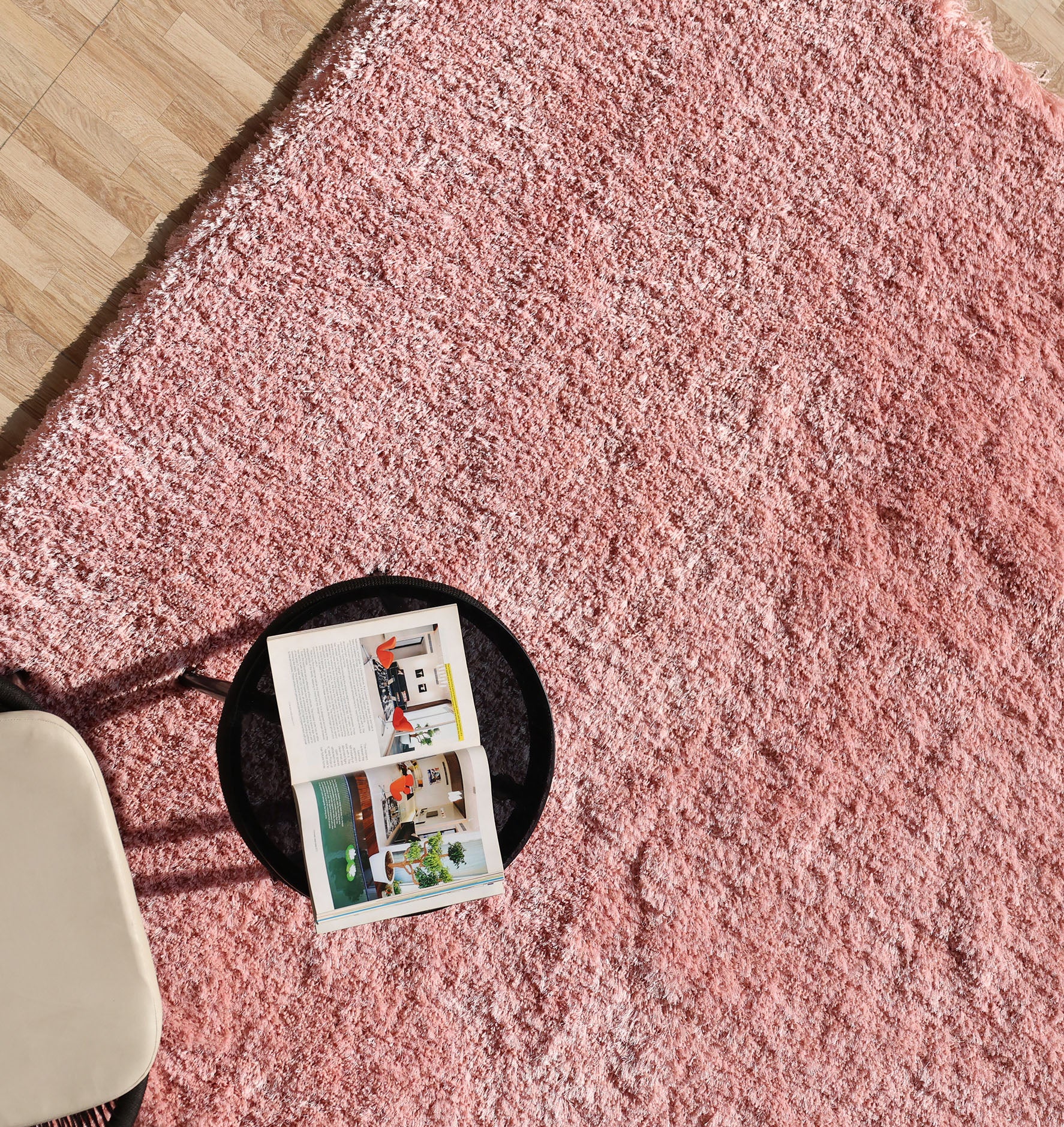 A round black tray with an open magazine sits on the Modern Ultra Soft Pink Color Shaggy Carpet for Bedroom (SG 042) by RAMSHA HOME, next to a beige cushioned seat atop a light wood floor.