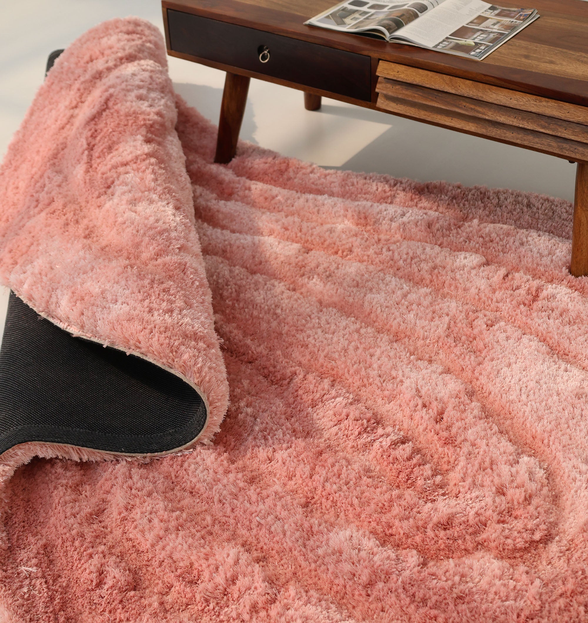 The Gold irregular Shaped Home Decor Shaggy Rug - SG 042 3D by RAMSHA HOME, with black non-slip backing partially flipped over, lies near a wooden table with an open magazine as sunlight streams onto the rug and floor.