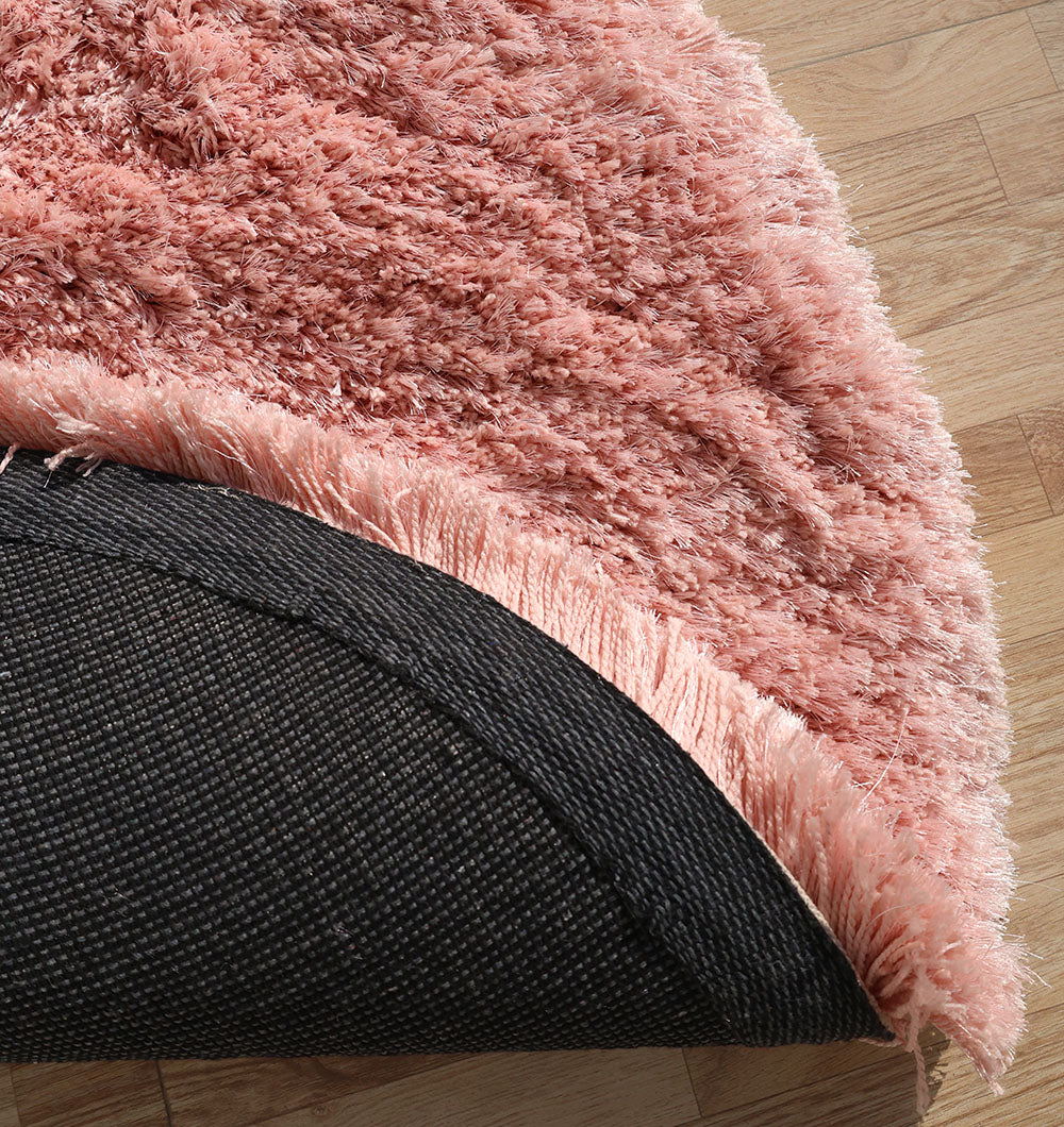 A close-up of the RAMSHA HOME Modern Ultra Soft Pink Shaggy Carpet for Bedroom (SG 042), showing its plush texture and a flipped corner revealing the black non-slip backing on a light wooden floor.