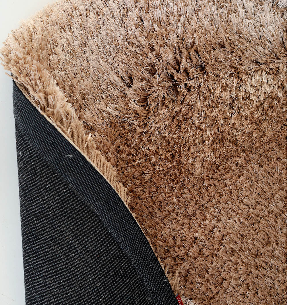 A close-up of the RAMSHA HOME Soft Plush Brown Shaggy Rug for Living Room - SG 043 shows one corner flipped, revealing a black textured backing and thick, ultra-soft beige fibers that look exceptionally inviting.