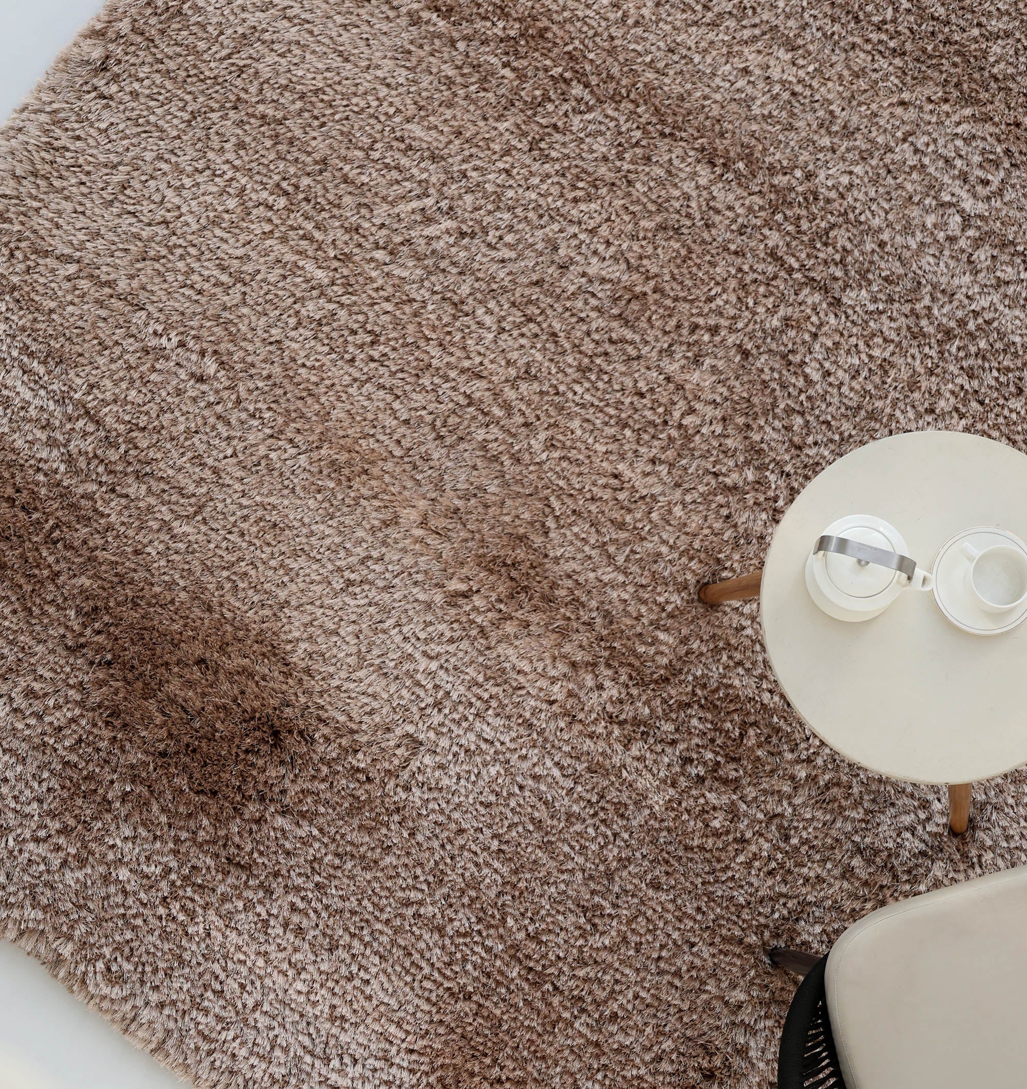 The RAMSHA HOME Soft Plush Brown Shaggy Rug for Living Room - SG 043 is shown from above, featuring darker patches, a round light table with a teapot and cup, and the edge of a chair in the lower right corner.