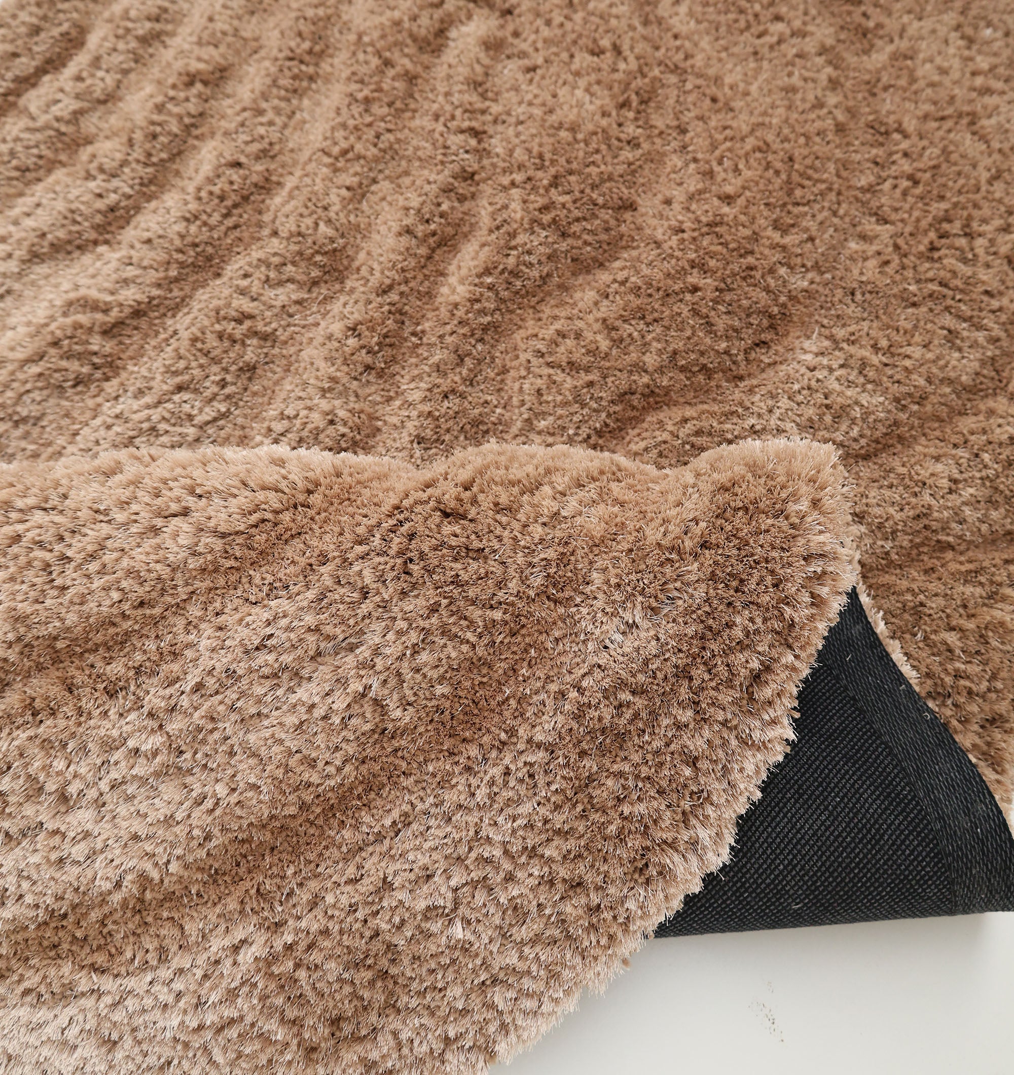 A close-up of the RAMSHA HOME Luxury Soft Touch Irregular Shaggy Area Rug - SG 043 3D shows its brown, fluffy texture and a lifted corner revealing black non-slip backing, displayed on a white surface.