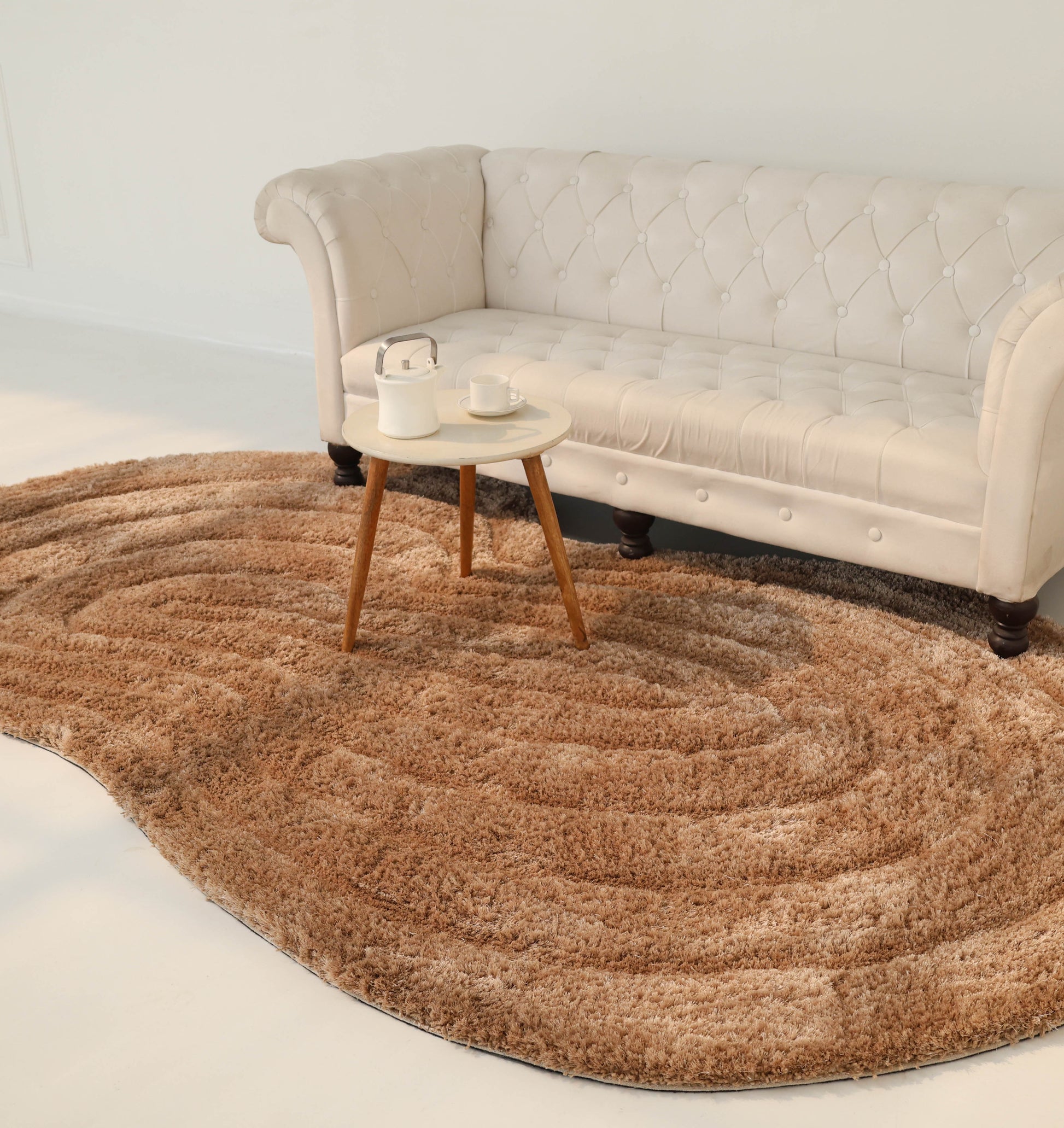 A cream-colored, tufted sofa sits against a white wall. In front, a small round table with two cups and a teapot rests on the RAMSHA HOME Luxury Soft Touch Irregular Shaggy Area Rug - SG 043 3D.