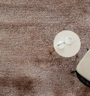 A small round table with a cup, saucer, and hourglass sits next to a light chair on the RAMSHA HOME Soft Plush Brown Shaggy Rug for Living Room (SG 043), viewed from above.