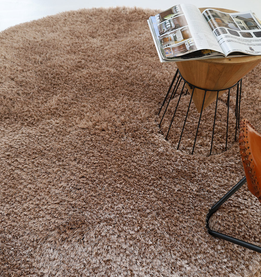 The RAMSHA HOME Soft Plush Brown Shaggy Rug for Living Room (SG 043) sits next to a brown leather chair with a metal frame, with a small wooden table holding an open magazine on the rug.